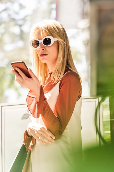 Belle fille blonde dans des lunettes de soleil tenant des sacs en papier et en utilisant un smartphone — Photo de stock