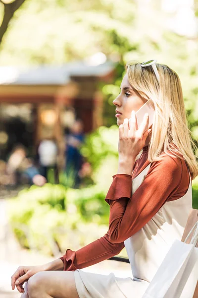 Вид сбоку красивой блондинки, говорящей по смартфону, сидя с сумками на лавочке — стоковое фото