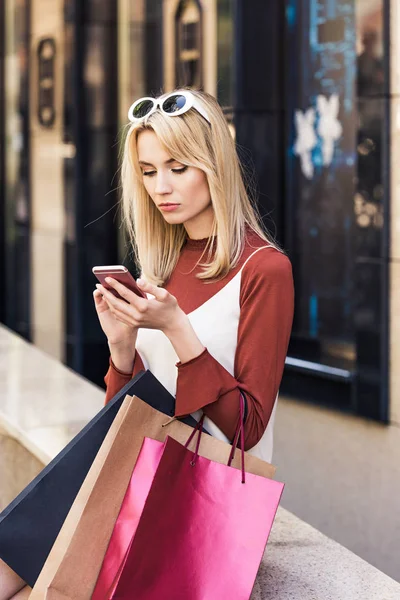 Красивая стильная молодая женщина, держащая сумки и используя смартфон — стоковое фото