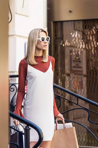 Belle fille blonde élégante dans des lunettes de soleil tenant des sacs à provisions et regardant loin dans la rue — Photo de stock