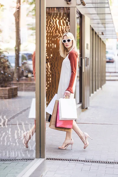 Вид сбоку стильной молодой женщины с сумками для покупок, смотрящей на камеру при входе в бутик — стоковое фото