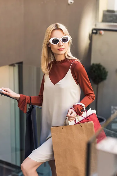 Selective focus of beautiful stylish blonde girl in sunglasses holding shopping bags on street — Stock Photo