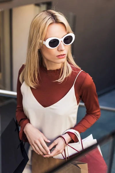 Blick aus der Vogelperspektive auf eine schöne junge Frau mit Sonnenbrille, die Papiertüten hält und wegschaut — Stockfoto