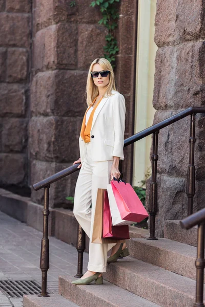 Stylish blonde woman in sunglasses holding shopping bags and looking at camera while standing on stairs — Stock Photo