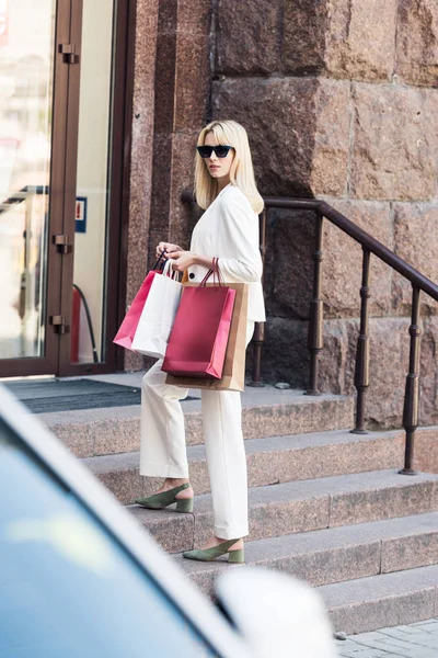 Bella donna bionda alla moda in possesso di borse della spesa e in piedi sulle scale — Foto stock