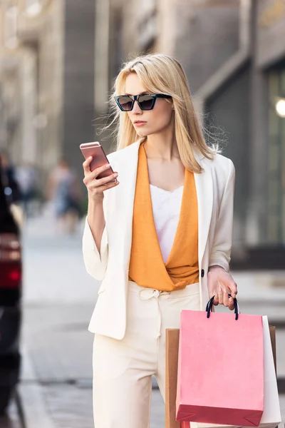 Mujer rubia con estilo en gafas de sol con bolsas de compras y el uso de teléfono inteligente en la calle - foto de stock