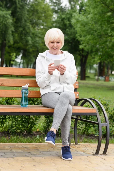 Пожилая женщина в спортивной одежде с помощью смартфона, сидя на скамейке со спортивной бутылкой воды — стоковое фото