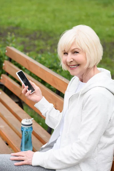 Anziana donna allegra utilizzando smartphone mentre seduto sulla panchina con bottiglia d'acqua sportiva — Foto stock