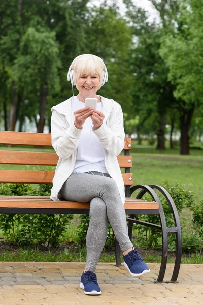 Alegre anciana escuchando música con auriculares y smartphone mientras está sentada en el banco en el parque - foto de stock