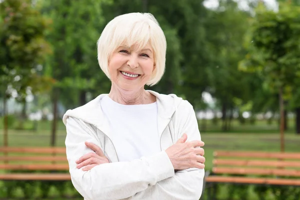 Smiling senior woman with crossed arms standing in park — Stock Photo