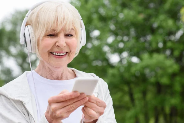 Donna anziana sorridente utilizzando smartphone e cuffie nel parco — Foto stock