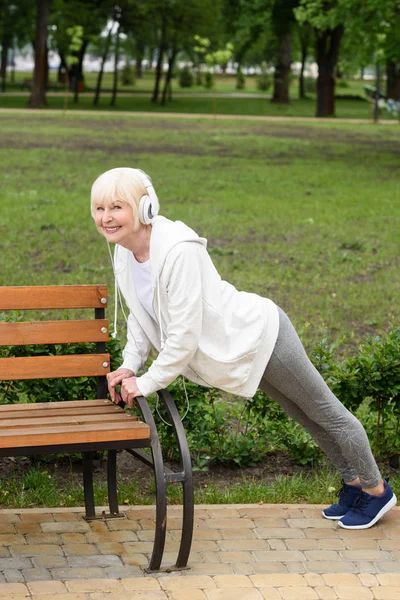 Старшая спортсменка в наушниках делает отжимания возле скамейки в парке — стоковое фото