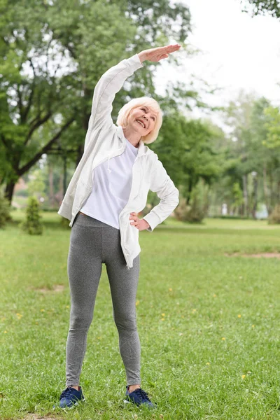 Elderly sportswoman stretching on green lawn in park — Stock Photo
