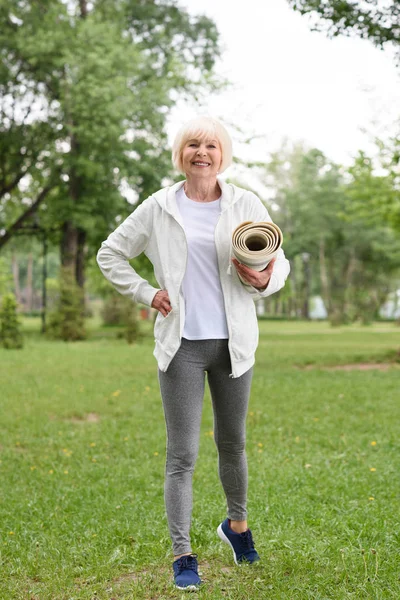 Усміхнена старша жінка стоїть з йога мат в зеленому парку — стокове фото