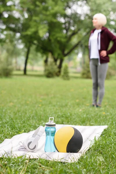 Selective focus of woman in park with medicine ball, water and headphones on yoga mat on foreground — Stock Photo