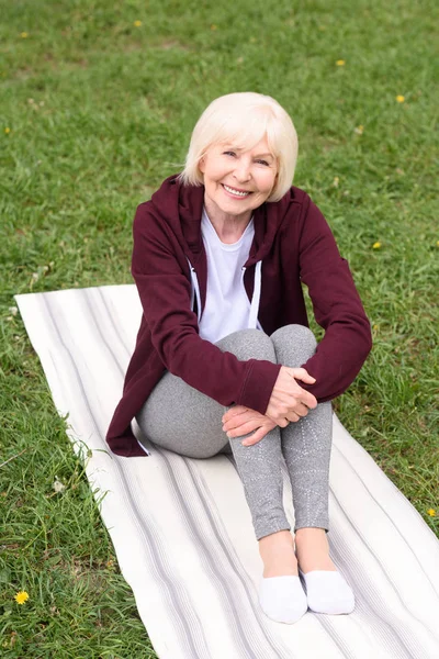 Старша жінка сидить на килимку для йоги на зеленому газоні — стокове фото