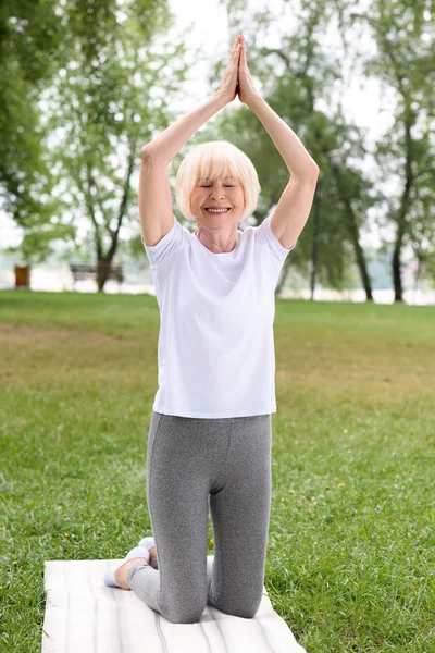 Пожилая женщина с жестами намасте практикующая йогу на коврике в парке — стоковое фото