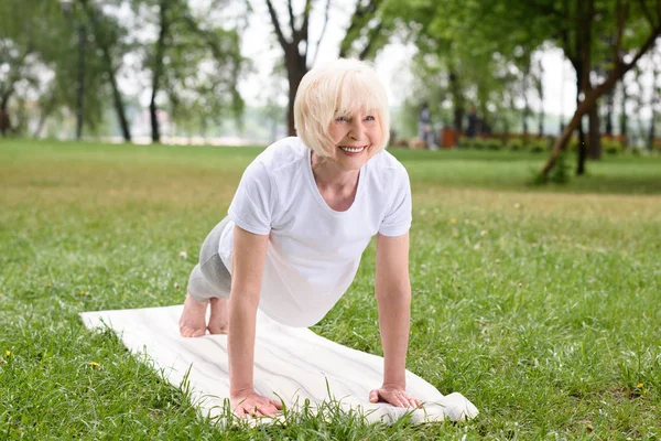 Anciana sonriente haciendo tablón en esterilla de yoga en el césped - foto de stock