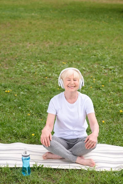 Happy senior woman listening music with headphones while sitting on yoga mat in park — Stock Photo