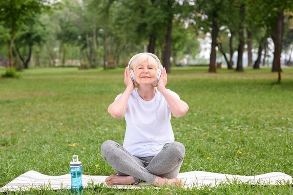 Happy elderly woman listening music with headphones while sitting on yoga mat with bottle of water in park — Stock Photo