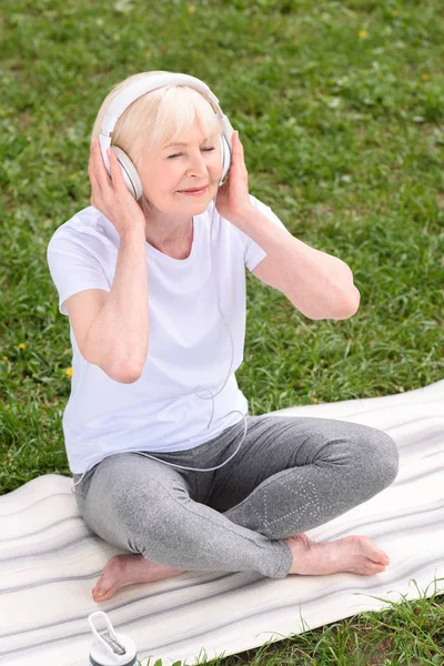 Senior woman listening music with headphones while sitting on yoga mat — Stock Photo
