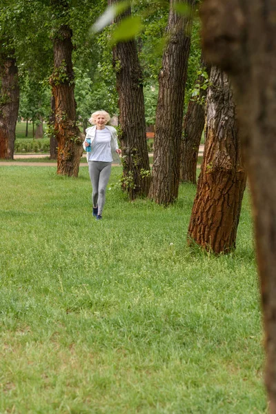 Elderly sportswoman running on green grass in park — Stock Photo