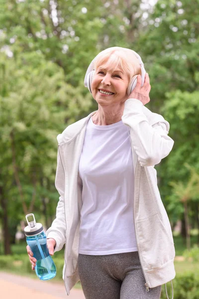 Happy senior sportswoman with water listening music with headphones in park — Stock Photo