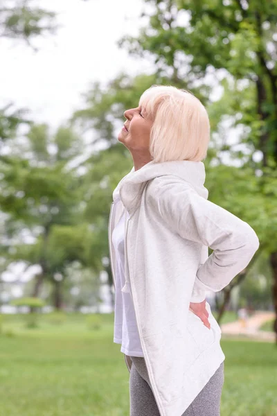 Caucasian elderly sportswoman with back ache standing in park — Stock Photo
