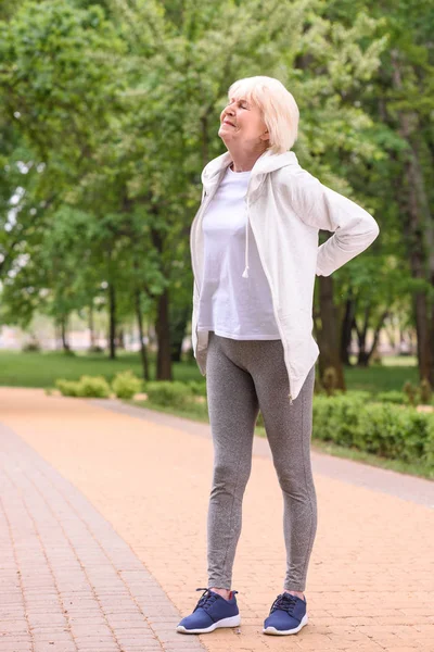 Elderly sportswoman with back ache standing in park — Stock Photo
