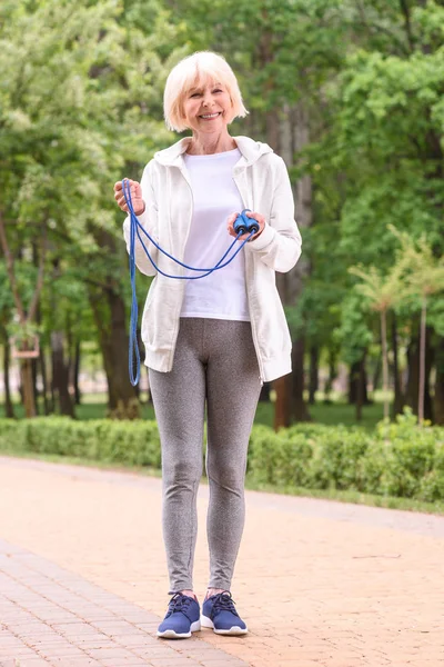 Cheerful elderly sportswoman standing with jump rope in park — Stock Photo