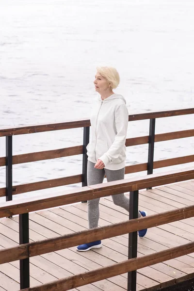 Senior woman with grey hair walking on wooden path near river — Stock Photo