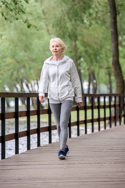 Mujer mayor en ropa deportiva caminando por el camino de madera en el parque cerca del río - foto de stock