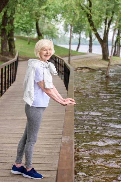 Happy senior sportswoman standing on wooden path near railings in park — Stock Photo