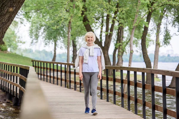 Mujer mayor en ropa deportiva caminando por el camino de madera en el parque - foto de stock