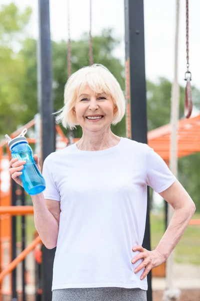 Sportiva anziana sorridente con acqua in bottiglia di sport in piedi su un campo sportivo — Foto stock