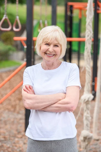 Sportiva anziana sorridente con le braccia incrociate in piedi su un campo sportivo — Foto stock