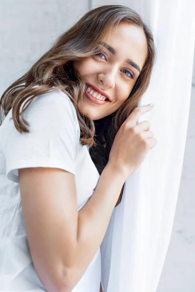 Vista de ángulo bajo de la hermosa mujer joven tocando la cortina blanca y sonriendo a la cámara - foto de stock