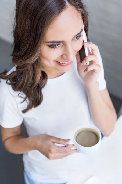 Vue grand angle de belle fille souriante tenant tasse de café et parlant par smartphone — Photo de stock