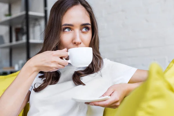 Hermosa chica morena pensativa bebiendo café y mirando hacia fuera en casa - foto de stock