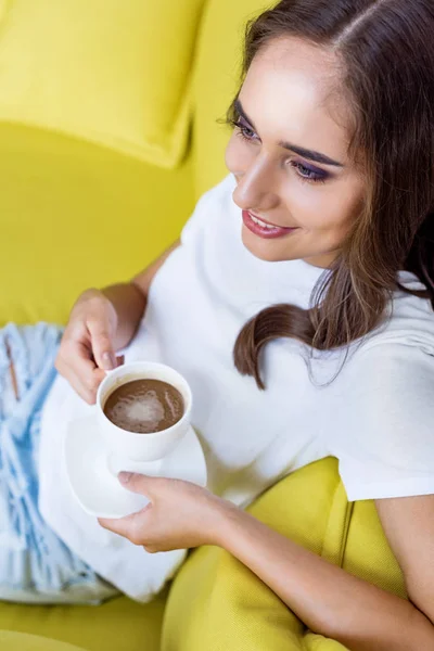 Високий кут зору красива усміхнена дівчина тримає чашку кави і сидить на дивані — стокове фото