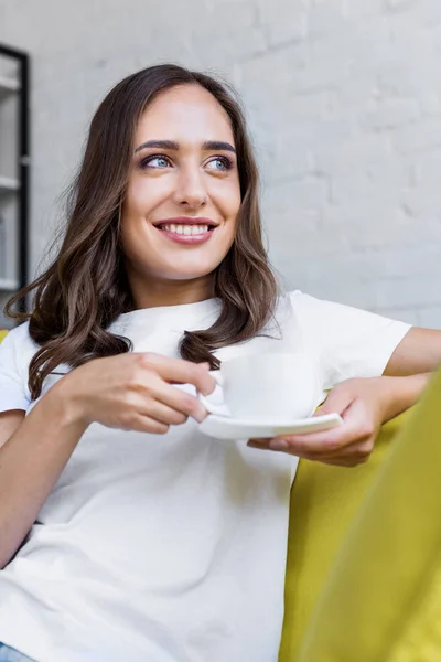 Heureuse jeune femme tenant une tasse de café et regardant loin à la maison — Photo de stock