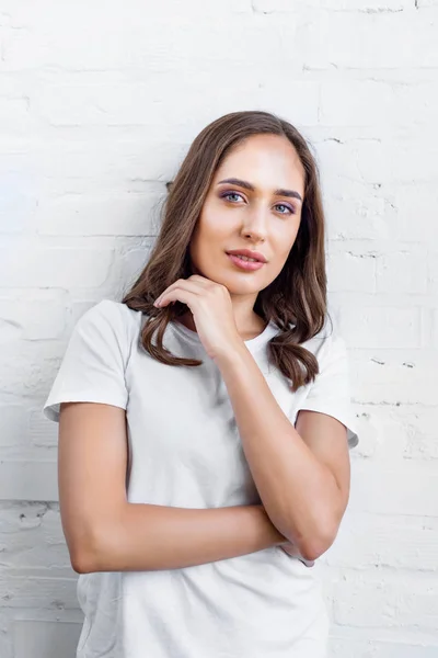 Belle jeune femme en t-shirt blanc debout près du mur de briques blanches et regardant la caméra — Photo de stock