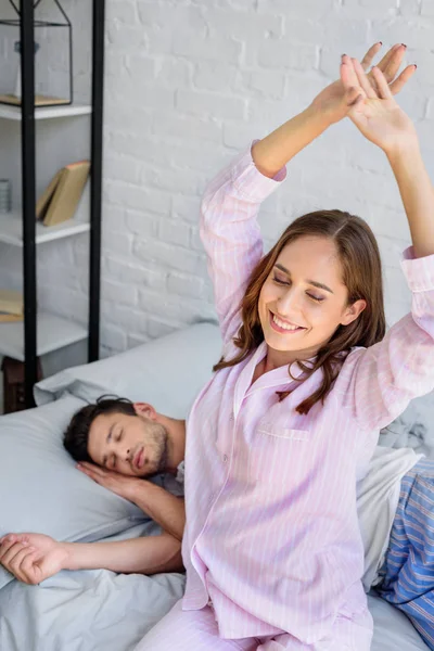 Happy young woman in pajamas stretching on bed while boyfriend sleeping behind — Stock Photo