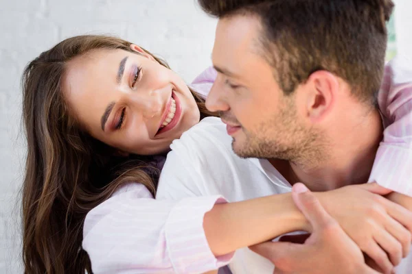 Feliz joven pareja en pijama sonriéndose y abrazándose por la mañana - foto de stock