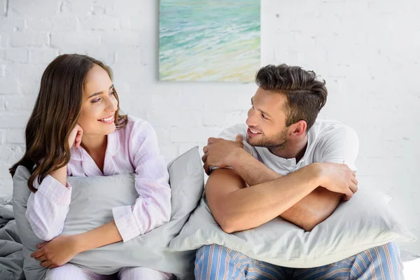 Happy couple in pajamas sitting on bed with pillows and looking at each other — Stock Photo
