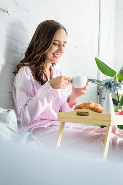 Belle femme souriante en pyjama rose buvant du café pour le petit déjeuner au lit — Photo de stock