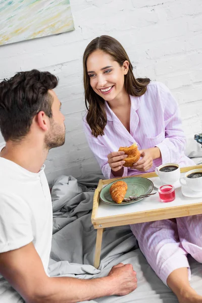 Joven riendo pareja en pijama desayunando con café y croissants en la cama juntos - foto de stock