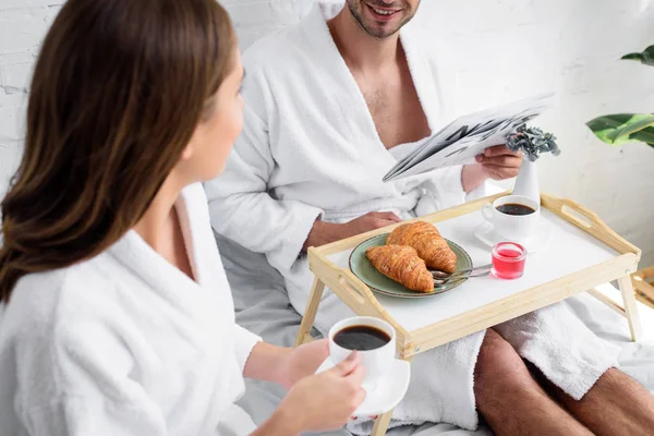 Joven esposa tomando café para el desayuno mientras marido leyendo periódico en la cama - foto de stock