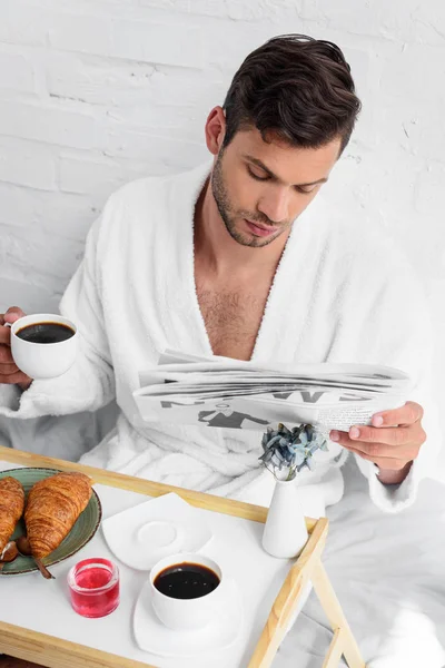 Joven hombre guapo en el periódico de lectura de albornoz durante el desayuno con café y cruasanes - foto de stock