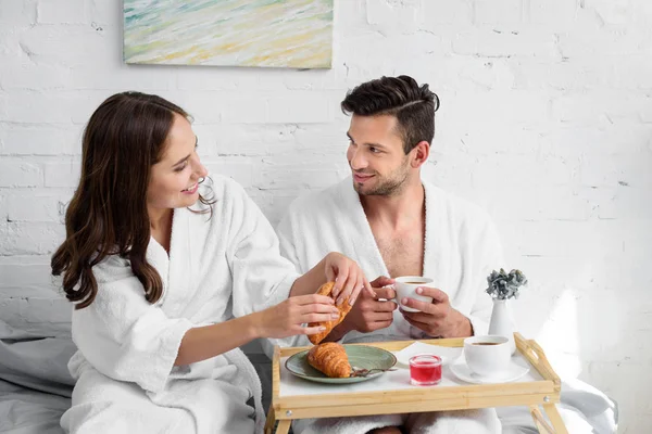 Jeune couple en peignoirs de bain ayant croissants et café pour le petit déjeuner au lit — Photo de stock
